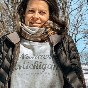 woman in a snowy landscape wearing a grey long sleeve Northern Michigan looks good on me shirt with forest green letters